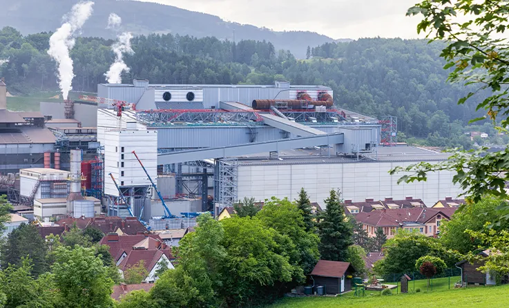 Industrieanlage mit rauchenden Schornsteinen in bewaldeter Hügellandschaft, im Vordergrund ein Wohngebiet (photo)